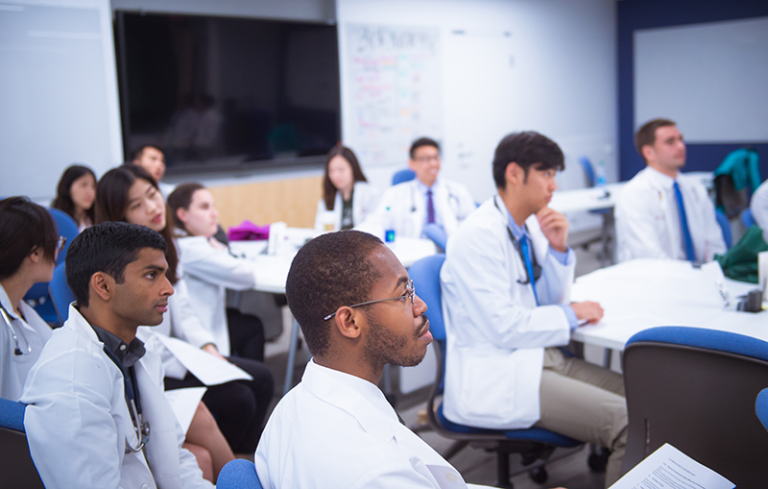 Students in white coats attend a clinical skills class.