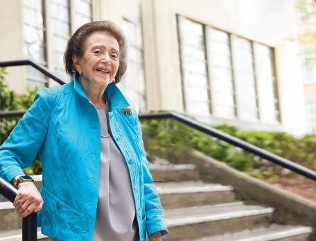 Nanette Wenger stands outside a beige building on the stairs. She smiles at the camera while wearing a bright blue suit jacket.