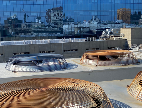 Large, flat fans rest on a rooftop. Behind the rooftop there is a building with glass walls reflecting the city skyline and blue sky.
