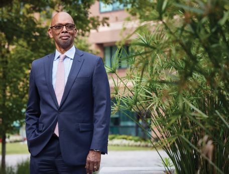 Herman Taylor stands in front of a brick building next to a large green plant. He is wearing a navy blue suit and light red tie, and smiling.