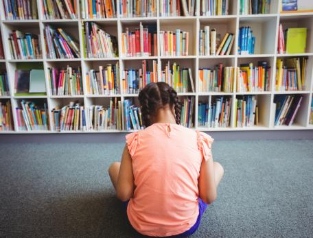 A girl in a pink shirt sits on the floor of a library facing the bookshelves, reading a book.