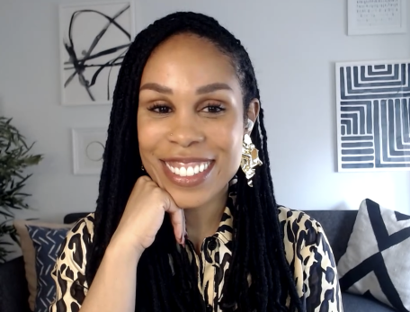 Uché Blackstock sits in a room with a white background, a blue couch and art on the walls. She looks at the camera.