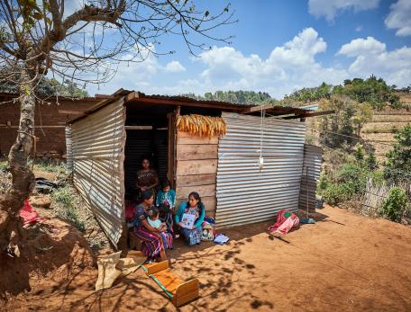A mother and children sit in the doorstep of a small shack in a rural area, speaking with a medical provider