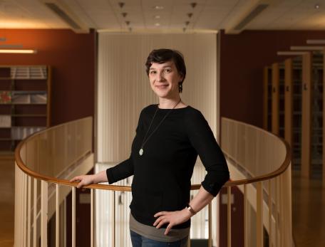 Talia Konkle poses by an indoor balcony in a library. She is wearing a black shirt and short dark hair and it looking at the camera.