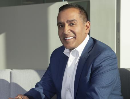 Sachin Jain sits on a grey couch wearing a blue suit. He looks at the camera and is smiling. 