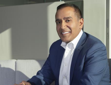 Sachin Jain sits on a grey couch wearing a blue suit. He looks at the camera and is smiling. 