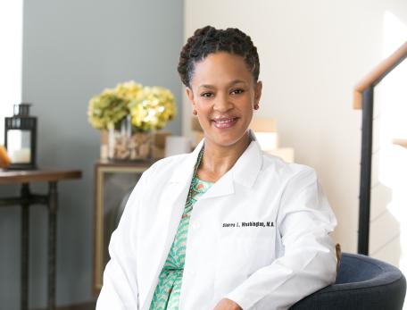 A woman in a white coat is seated on a chair in an office, smiling at the camera