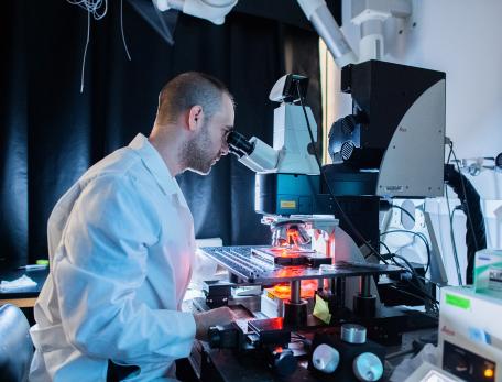A scientist wearing a white lab coat looks into a high-powered microscope in a dimly lit laboratory, surrounded by lab equipment.
