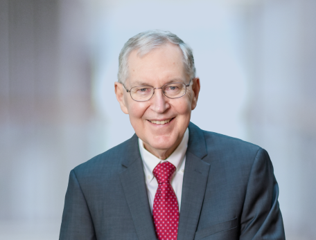 A man in a red tie and black suit jacket looks at the camera smiling with hands in his pockets