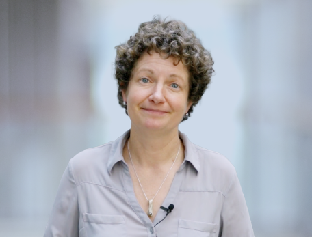 A woman with short curly hair in a grey button down shirt looks at the camera