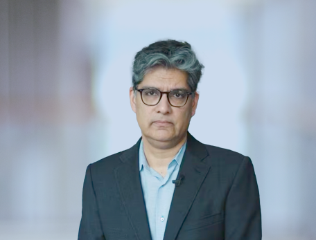 A man in a suit jacket and blue button down shirt with glasses stares at the camera with a serious expression