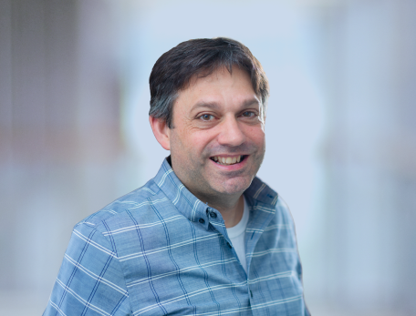 A man with brown hair in a checkered button down shirt looks at the camera against a neutral background