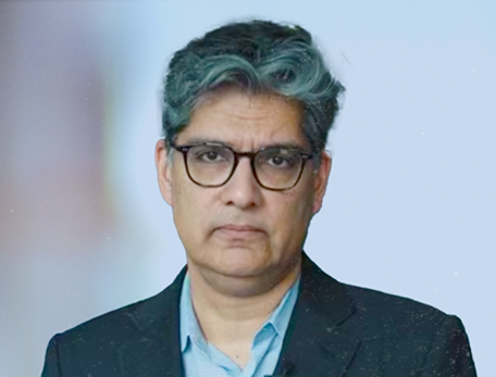 A man in a suit jacket and blue button down shirt with glasses stares at the camera with a serious expression