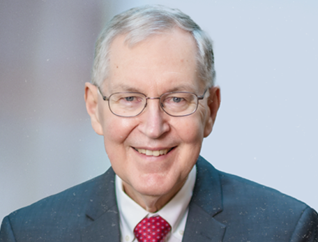 A man in a red tie and black suit jacket looks at the camera smiling with hands in his pockets