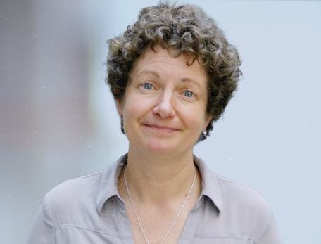 A woman with short curly hair in a grey button down shirt looks at the camera