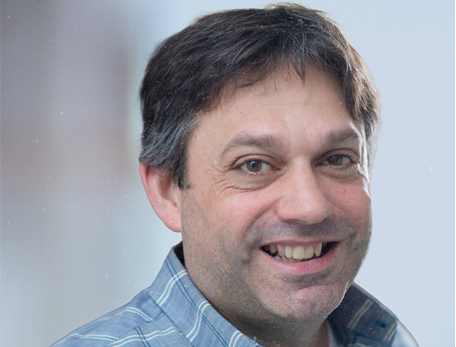 A man with brown hair in a checkered button down shirt looks at the camera against a neutral background