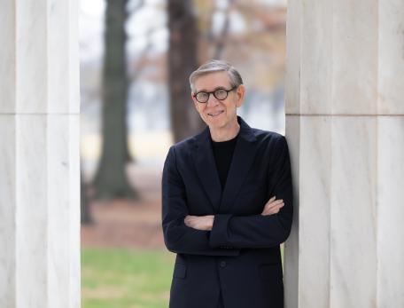 A man in a black shirt and suit jacket with thick rimmed glasses leans against a marble column outside with arms crossed and smiles slightly at the camera