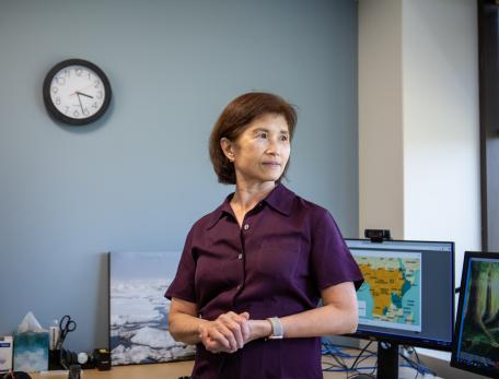 A woman stands in an office in front of two computer screens and looks toward the window with a serious expression.