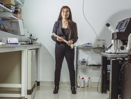 A woman stands in a laboratory holding a femur bone, surrounded by scientific equipment and lab supplies