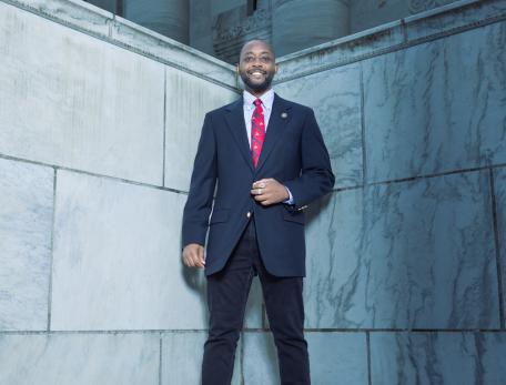 A man in a formal suit and red tie stands confidently on a marble ledge outside a grand building with tall columns, conveying professionalism and poise.