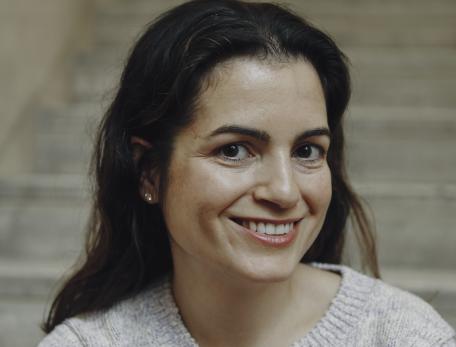Smiling woman with dark hair wearing a light sweater, sitting on stone steps. The background is softly blurred, conveying a warm and inviting atmosphere.