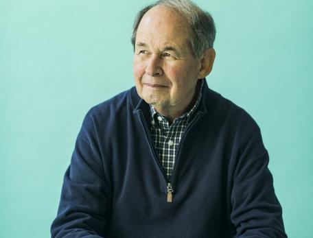 A man in a navy sweater sits at a teal table with a thoughtful expression. A cup of coffee is placed in front of him, against a teal background.