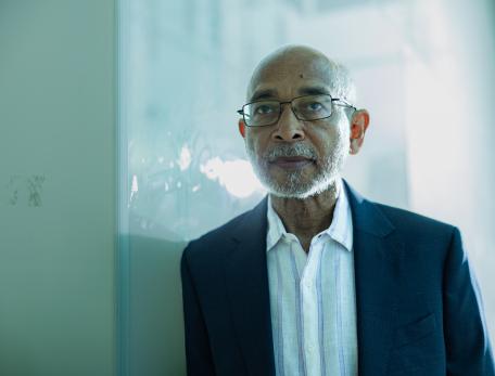 A man in glasses and a navy blazer stands pensively against a reflective glass wall, soft natural light highlighting his thoughtful expression.