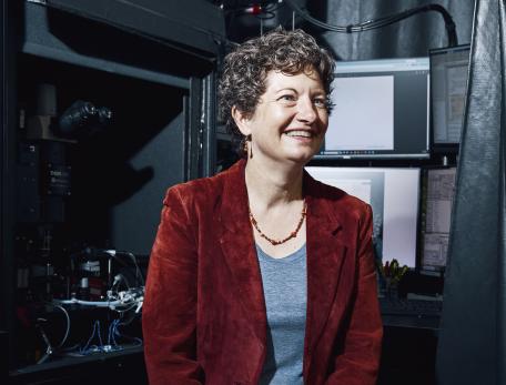 A woman with short curly hair, wearing a red blazer and jeans, sits on a stool in a lab with computers and scientific equipment in the background. She has a wide smile and is looking off to the side.