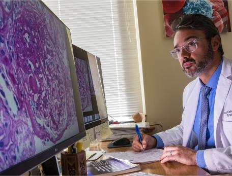 A doctor in a lab coat looks intently at two computer monitors displaying microscopic images, while taking notes at his desk.
