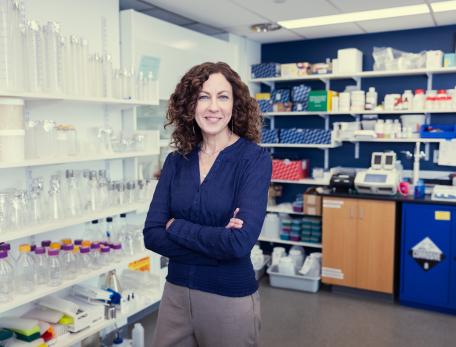 Smiling woman standing with folded arms in a laboratory surrounded by glassware, scientific supplies, and equipment.