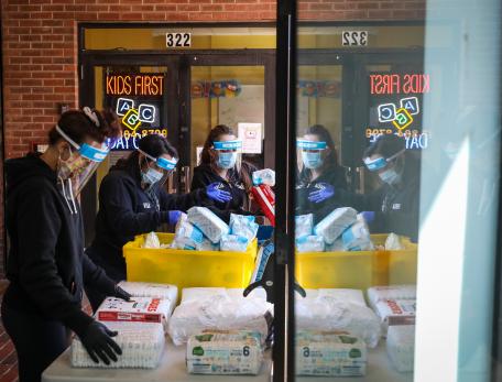 Three people wearing protective masks organize food, diapers, and baby formula on a table.