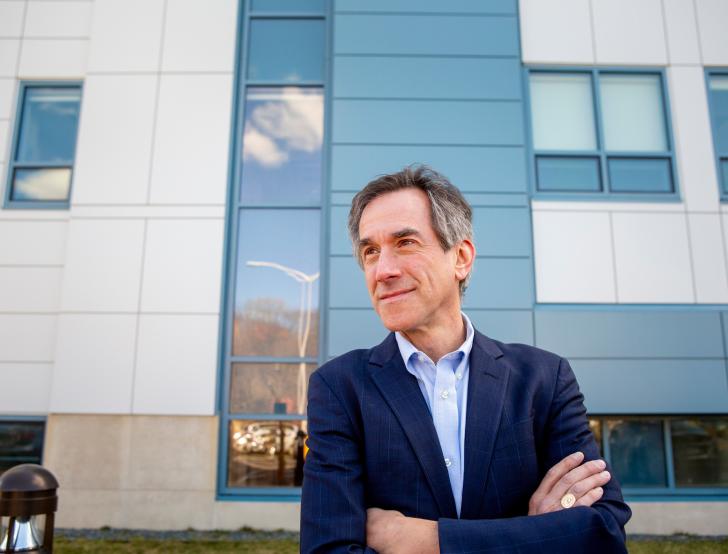 Andrew Budson stands in front of a blue and white building and looks off into the distance with his arms crossed