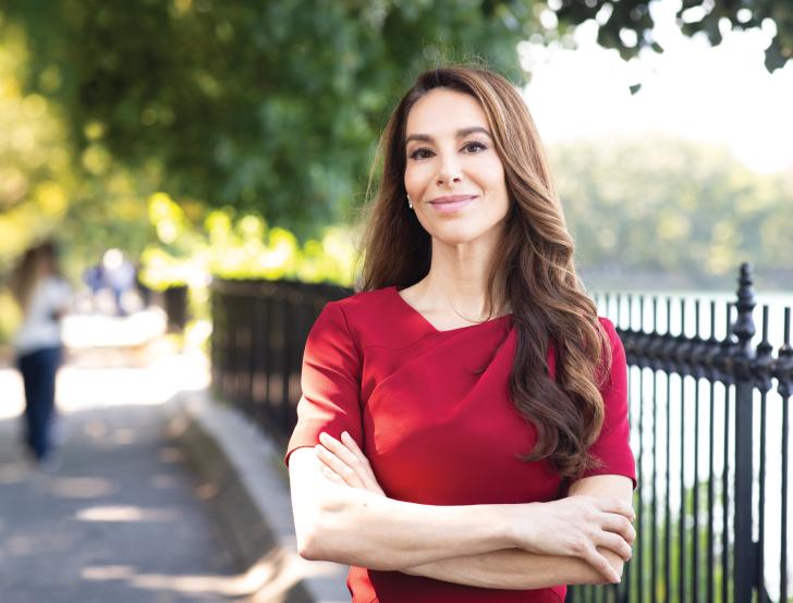 A woman with long brown hair and a red shirt stands outside on a sidewalk with arms crossed, smiling at camera 