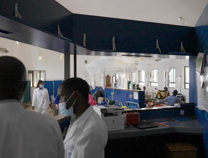 Interior view of a hospital ward with patients resting in beds separated by blue partitions and hanging white mosquito nets. In the foreground, health care workers wearing white coats and face masks stand near a reception desk .