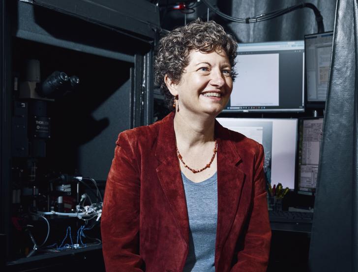 A woman with short curly hair, wearing a red blazer and jeans, sits on a stool in a lab with computers and scientific equipment in the background. She has a wide smile and is looking off to the side.