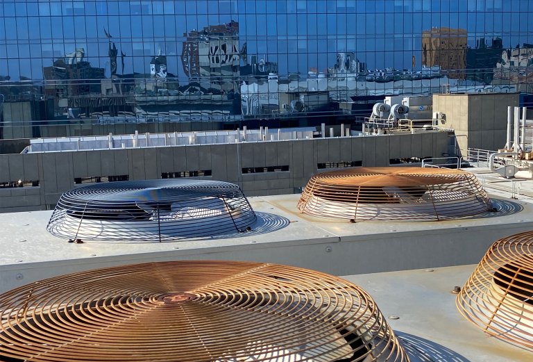 Large, flat fans rest on a rooftop. Behind the rooftop there is a building with glass walls reflecting the city skyline and blue sky.