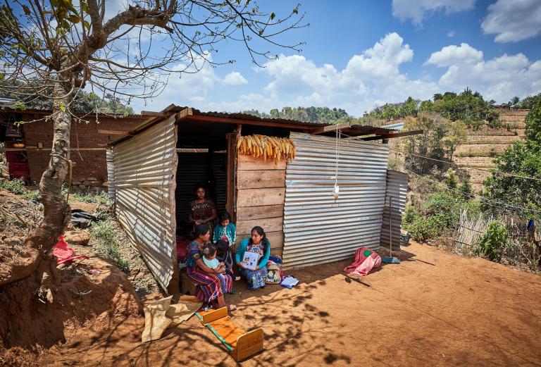 A mother and children sit in the doorstep of a small shack in a rural area, speaking with a medical provider
