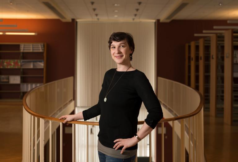 Talia Konkle poses by an indoor balcony in a library. She is wearing a black shirt and short dark hair and it looking at the camera.