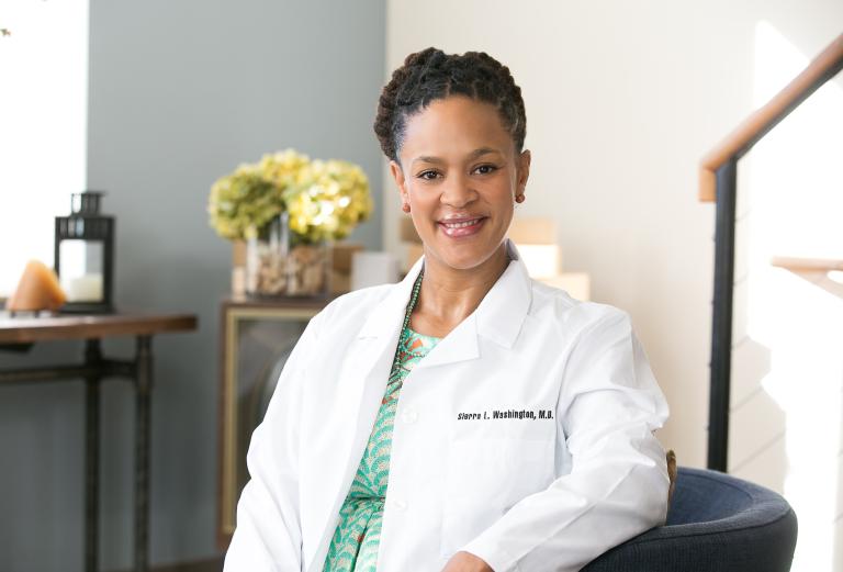 A woman in a white coat is seated on a chair in an office, smiling at the camera