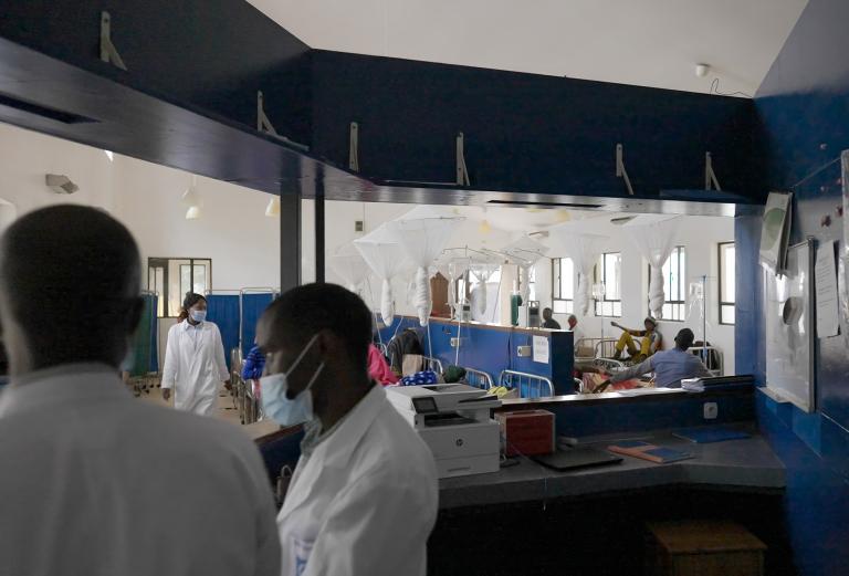 Interior view of a hospital ward with patients resting in beds separated by blue partitions and hanging white mosquito nets. In the foreground, health care workers wearing white coats and face masks stand near a reception desk .