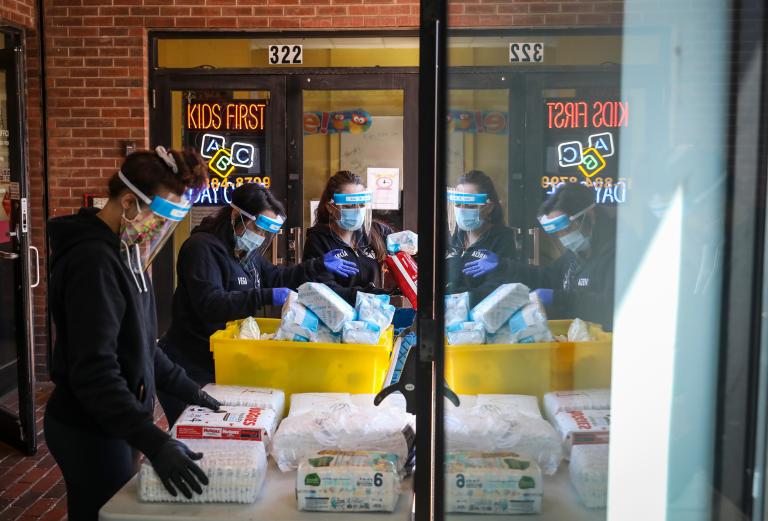 Three people wearing protective masks organize food, diapers, and baby formula on a table.