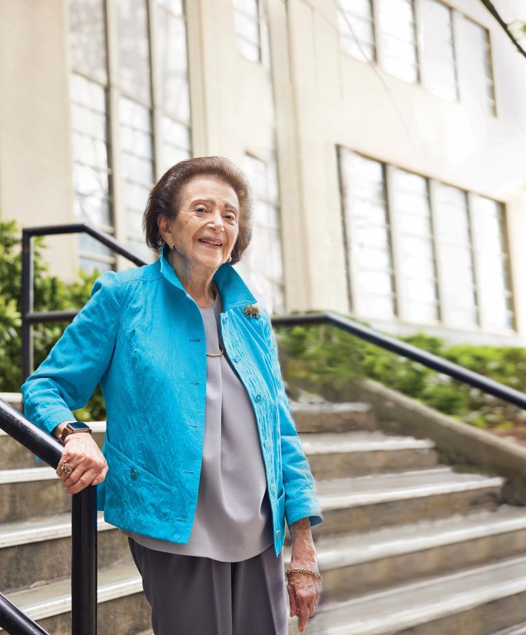 Nanette Wenger stands outside a beige building on the stairs. She smiles at the camera while wearing a bright blue suit jacket.