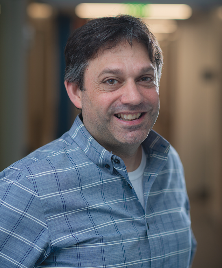 Stephen Liberles stands in a hallway and looks at the camera, smiling, wearing a blue checkered shirt