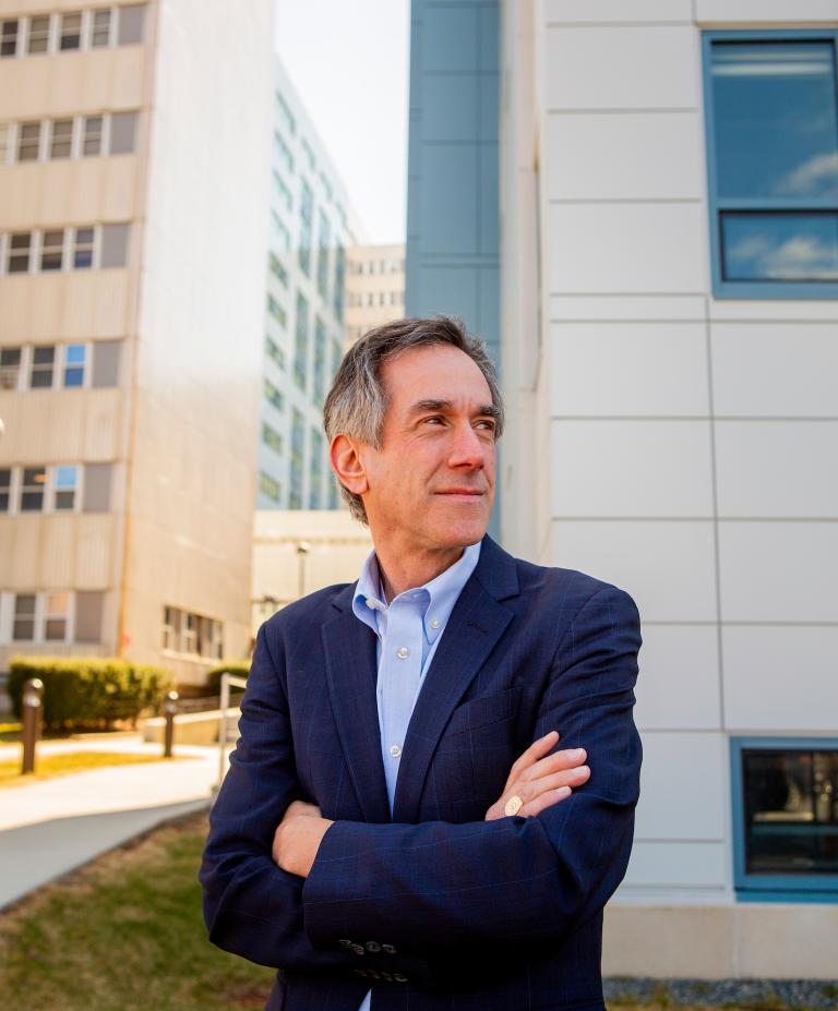 Andrew Budson stands in front of a blue and white building and looks off into the horizon with his arms crossed