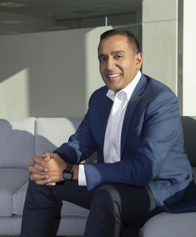 Sachin Jain sits on a grey couch wearing a blue suit. He looks at the camera and is smiling. 