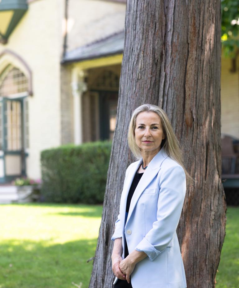 A woman in a white suit jacket stands in front of a large pine tree trunk in front of a light yellow building