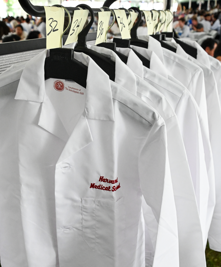 Several white coats embroidered with "Harvard Medical School" hanging on a hangers on a rack on white coat day
