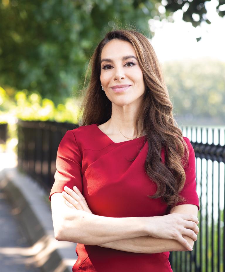 A woman with long brown hair and a red shirt stands outside on a sidewalk with arms crossed, smiling at camera