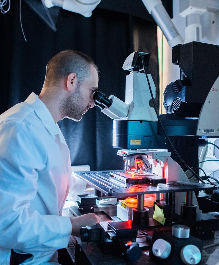 A scientist wearing a white lab coat looks into a high-powered microscope in a dimly lit laboratory, surrounded by lab equipment.