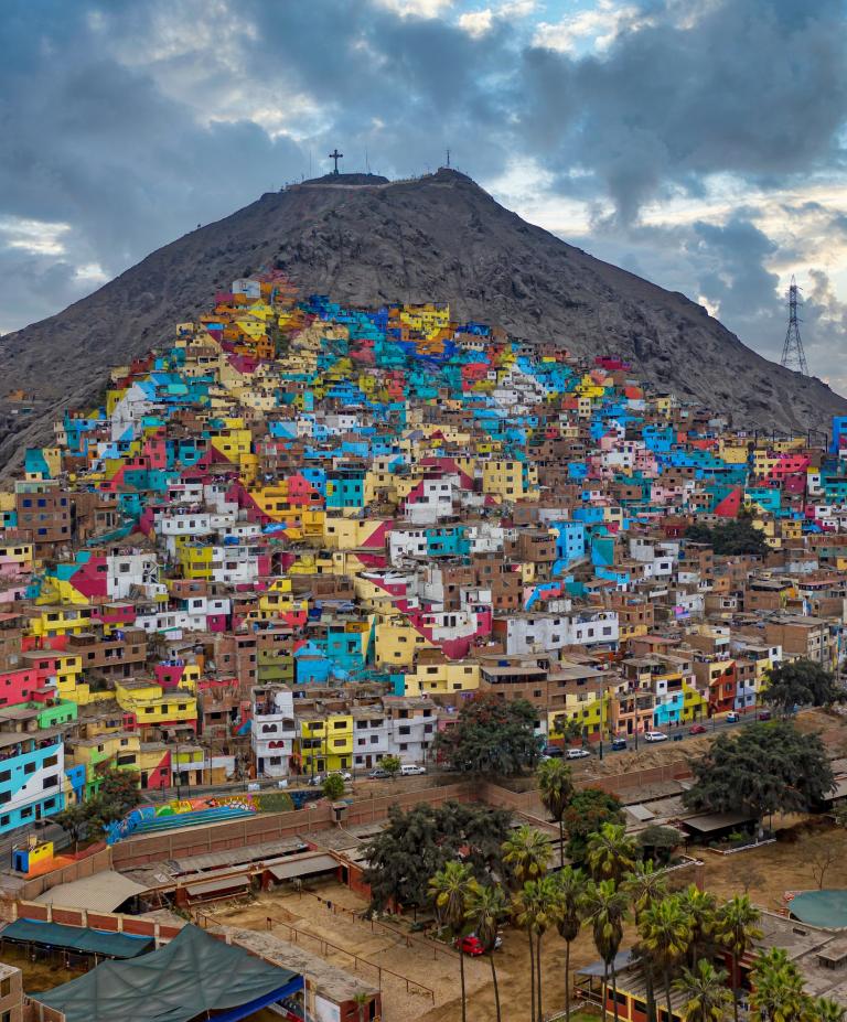 Aerial view of San Cristóbal Hill in the Rímac district of Lima, Peru, densely packed with informal housing and winding, narrow streets. The hillside is covered with colorful, tightly clustered homes built into the steep terrain, illustrating the crowded urban development of the area.